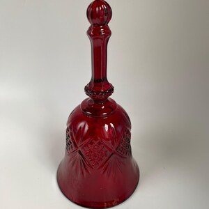 May include: A deep red glass bell featuring a pressed pattern of diamond shapes and radiating lines on the flared body. The handle is slender with a spherical top and a decorative collar where it connects to the main body of the bell.