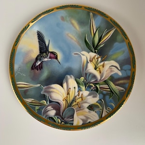 May include: Decorative plate featuring a hummingbird in flight near white lilies. The plate has a gold rim with a green border. The background is a soft blue with yellow highlights. A beautiful piece of art.