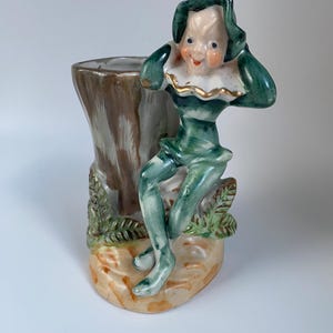 May include: A ceramic figurine vase featuring a green-clad elf sitting on a tree stump. The elf wears a pointed hat and a white ruffled collar with gold trim. The base is tan with green leaf accents, and the hollow tree trunk serves as a vessel.