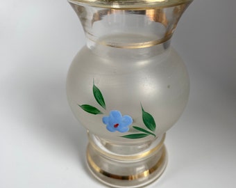 May include: A small, clear glass vase with a frosted body and a gold rim. The vase features a painted blue flower with green leaves. The base and the top rim are accented with gold trim. The vase is approximately 5 inches tall.