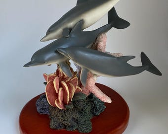 May include: A sculpture of three gray dolphins leaping above coral and seaweed, mounted on a reddish-brown oval base. The dolphins have white bellies and are positioned to appear as if they are swimming. The coral is pink and the seaweed is dark green.