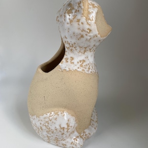 May include: A ceramic cat-shaped planter with a speckled white and beige glaze. The cat is sitting upright with its head turned to the side, featuring a hollowed-out area for planting. The planter has a rustic, handmade appearance.