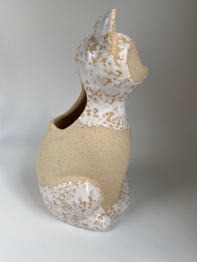 May include: A ceramic cat-shaped planter with a speckled white and beige glaze. The cat is sitting upright with its head turned to the side, featuring a hollowed-out area for planting. The planter has a rustic, handmade appearance.