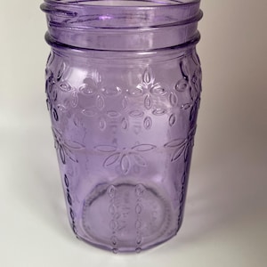 May include: A purple glass jar with a decorative floral pattern. The jar has a wide mouth and a screw-on rim. The glass is a translucent purple color. The jar is empty and sits on a white surface.