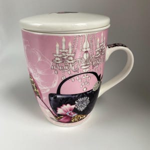 May include: A pink ceramic teacup with a lid, featuring a black handbag, a pair of high heels, and a chandelier with crystals. The teacup has a handle and a floral pattern.