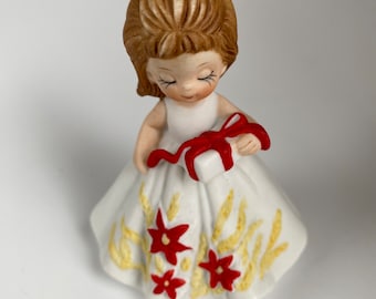 Könnte beinhalten: Keramikfigur eines Mädchens in einem weißen Kleid mit roten floralen Akzenten und einer roten Schleife im Haar. Sie hält ein kleines Geschenk mit einem roten Band. Die Figur hat geschlossene Augen und einen ruhigen Ausdruck.