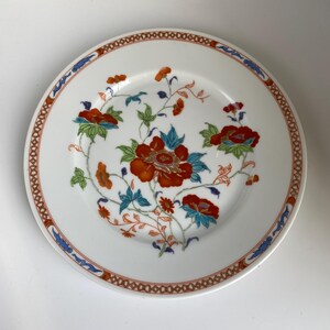 May include: White ceramic plate with a floral design. The plate features a red, orange, and blue floral pattern in the center, with a decorative border of red and blue accents. The plate is round and has a classic design.