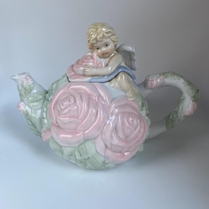May include: A decorative porcelain teapot featuring a cherub figurine atop the lid. The teapot is adorned with large pink roses and green leaves. The handle and spout are also decorated with floral accents. The cherub has blonde hair and wears a blue sash.