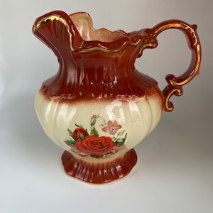 May include: A brown and white ceramic pitcher with a gold rim and handle. The pitcher features a floral design with red roses and pink flowers.