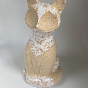 May include: A ceramic cat ornament in a seated pose. The cat features a speckled beige body with white detailing on the face, chest, and tail. The ears are pointed, and the eyes are not visible. The ornament is set against a white backdrop.