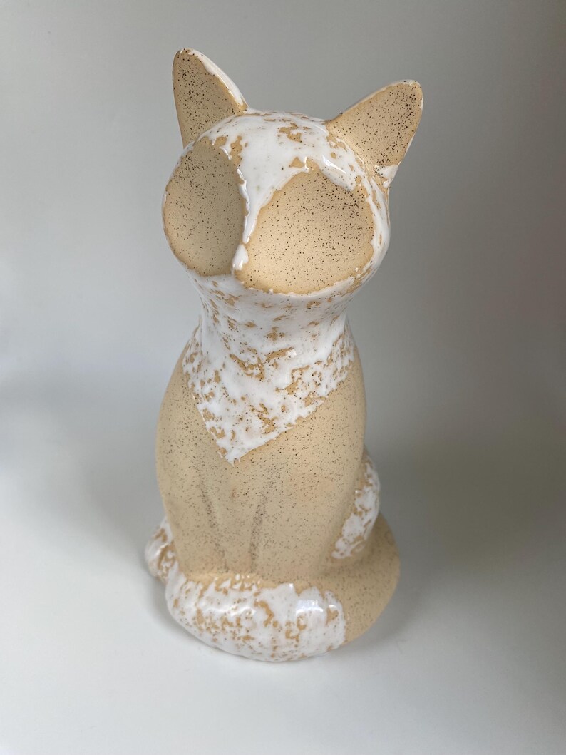 May include: A ceramic cat ornament in a seated pose. The cat features a speckled beige body with white detailing on the face, chest, and tail. The ears are pointed, and the eyes are not visible. The ornament is set against a white backdrop.