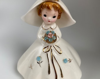 Pode incluir: Uma figurinha de porcelana branca de uma noiva vestindo um vestido branco com véu e segurando um buquê de flores azuis. A figurinha tem detalhes dourados e flores azuis no vestido.