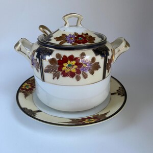May include: A ceramic sugar bowl with a lid and two handles, accompanied by a matching saucer and a small spoon. The set features a cream-colored base with hand-painted floral motifs in red, purple, and yellow, accented by dark blue and gold trim.