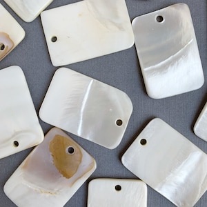 May include: A collection of eight white rectangular mother of pearl shell tags with a hole in the top. The tags have a smooth, glossy finish and a subtle iridescent sheen.