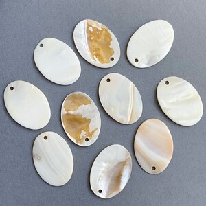 May include: Ten oval-shaped mother of pearl shell embellishments with a hole in the top. The shells are white with brown and gold iridescent patterns.
