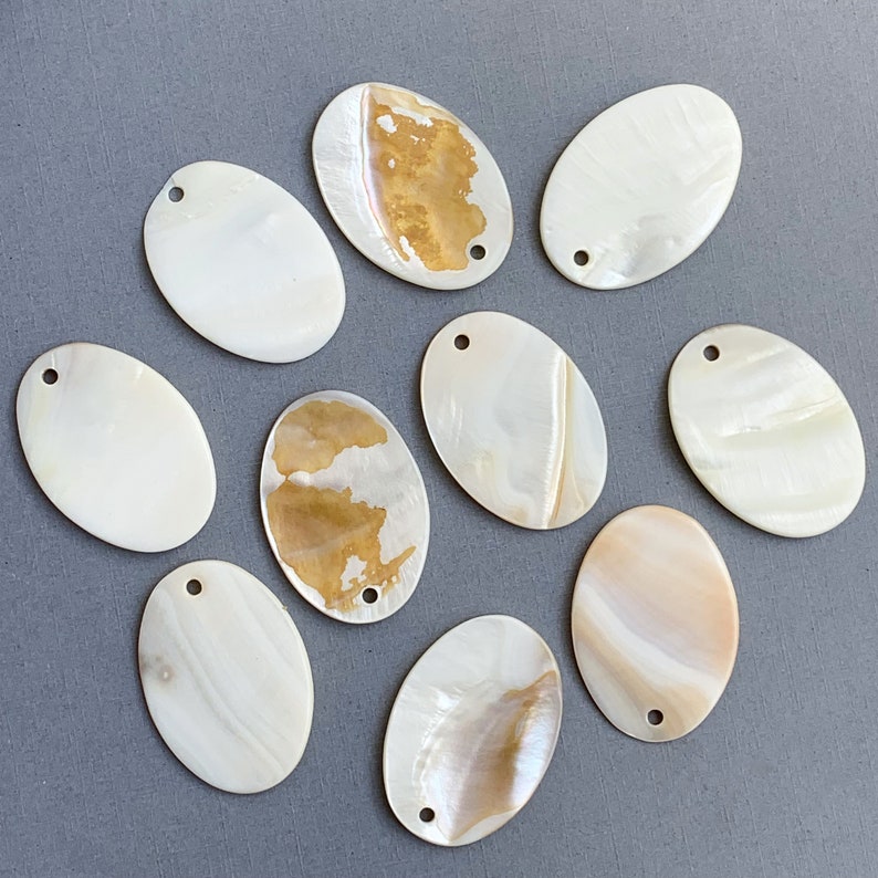 May include: Ten oval-shaped mother of pearl shell embellishments with a hole in the top. The shells are white with brown and gold iridescent patterns.