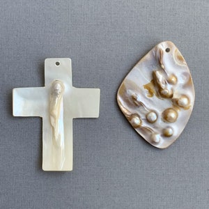 May include: Two iridescent shell pendants. One is a white cross with a textured center. The other is a teardrop shape with a pearl-like texture and several raised, pearl-like accents. Both have small holes for stringing.