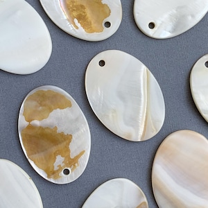 May include: A collection of oval-shaped, white mother-of-pearl shell pendants with a hole drilled at the top. The shells have a natural iridescent sheen and some have a light brown or gold color.