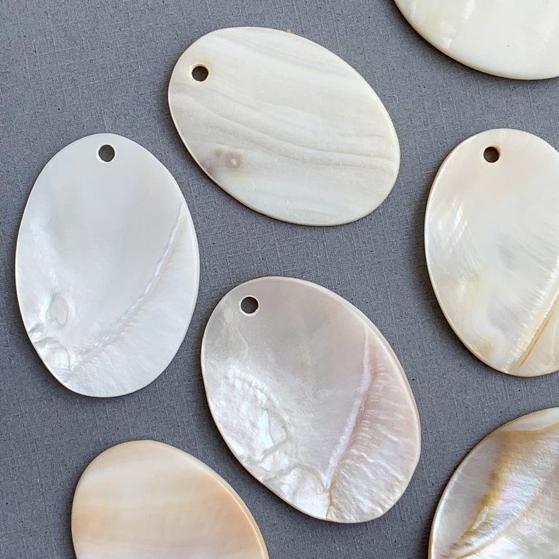 May include: Six oval-shaped mother of pearl shell pendants with a hole drilled at the top. The shells are a creamy white color with iridescent highlights.