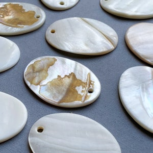 May include: A collection of oval-shaped, white mother-of-pearl shell pendants with a hole drilled at the top. Some of the shells have a brown, iridescent pattern.