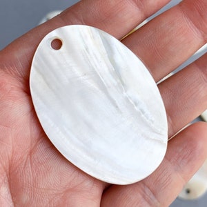May include: A white oval-shaped mother of pearl shell with a small hole drilled in the top. The shell has a smooth, iridescent surface.