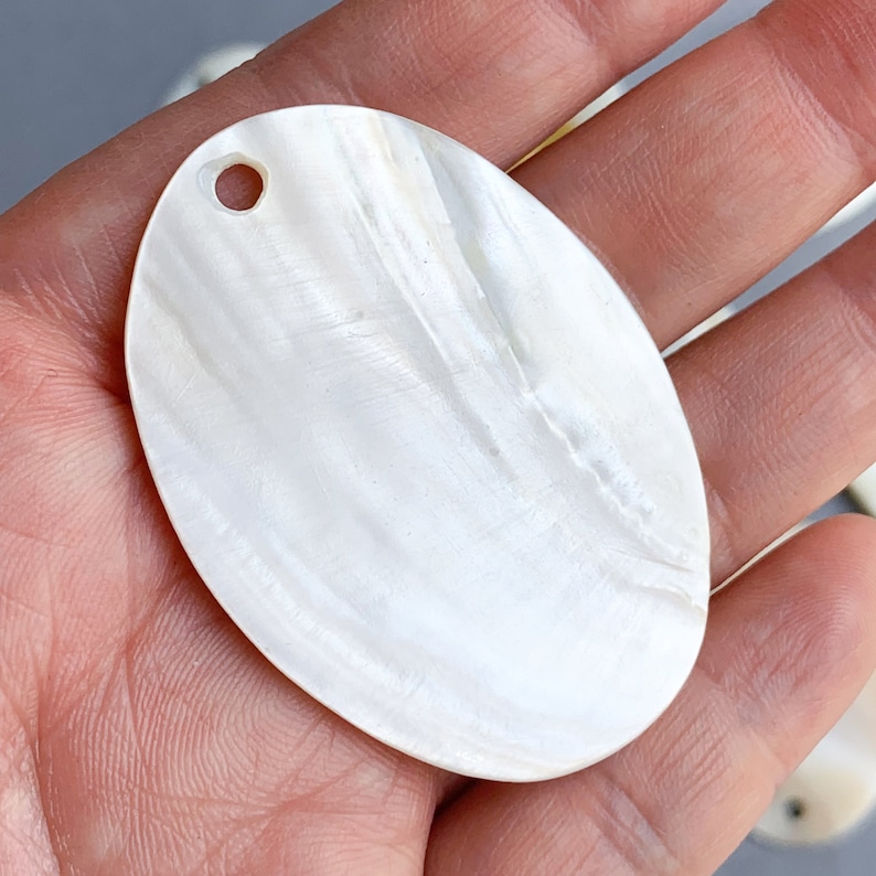 May include: A white oval-shaped mother of pearl shell with a small hole drilled in the top. The shell has a smooth, iridescent surface.
