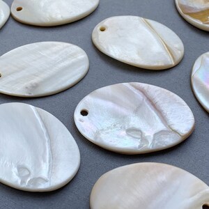 May include: Oval shaped mother of pearl shell with a hole drilled at the top. The shell has a white iridescent finish with a subtle pearlescent sheen.
