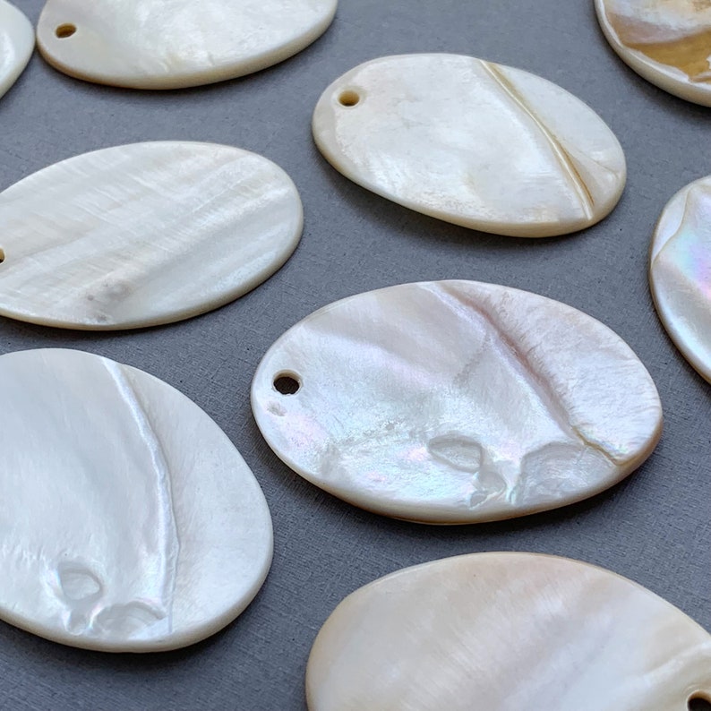 May include: Oval shaped mother of pearl shell with a hole drilled at the top. The shell has a white iridescent finish with a subtle pearlescent sheen.