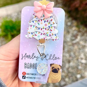 May include: A white Christmas tree with pink bow and gold star on top, with colorful lights and snowflakes. The tree is attached to a clear badge reel with a sparkly silver charm. The badge reel is on a card with the text "Harley & Chloe Badge Reels" and social media handles.