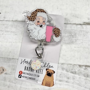 May include: A clear plastic badge reel with a Santa Claus design. Santa is wearing a leopard print hat and holding a pink cup. The badge reel has a white glitter charm with the text "Harley and Chloe Badge Reel" and a website address.