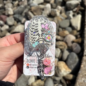May include: A clear badge reel with a cartoon rib cage design and colourful internal organs. The badge reel has a silver skeleton charm and a pink glitter accent. The text "Harley & Chloe" and "Badge Reel" are visible.