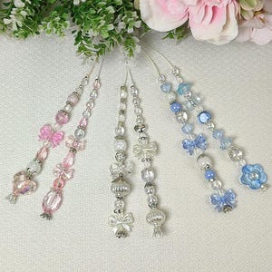 Puede incluir: Varios adornos de cuentas en tonos rosa, transparente y azul. Cada adorno presenta una variedad de cuentas, incluyendo cuentas transparentes, iridiscentes y perladas, con detalles de lazo y flor. Los adornos cuelgan sobre un fondo blanco.