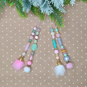 Może przedstawiać: Dwa różowe i zielone breloczki z koralików z białymi i różowymi puszystymi frędzlami wiszące na brązowym tle w białe kropki.