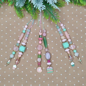 Könnte beinhalten: Dekorative Perlenornamente in Pastellfarben mit rosa, mintgrünen und goldenen Akzenten. Jedes Ornament hat ein einzigartiges Design mit Perlen, Charms und einem Cupcake- oder Eiswaffel-Charm. Die Ornamente hängen an einem Blumenarrangement.