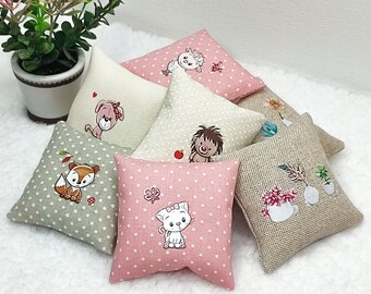 Könnte beinhalten: Sechs kleine Dekokissen mit verschiedenen Tier- und Blumenmotiven. Die Kissen sind aus einem weichen, hellen Stoff mit einem dezenten Punkte-Muster gefertigt. Die Designs beinhalten einen Fuchs, eine Katze, einen Bären, einen Igel, ein Schaf und ein Blumenarrangement.