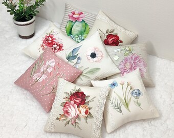 Könnte beinhalten: Eine Sammlung von acht dekorativen Kissen mit floralen und botanischen Designs. Die Kissen sind aus einem weichen, strukturierten Stoff gefertigt und in verschiedenen Farben gehalten, darunter Rosa, Weiß, Grün und Rot. Die Kissen sind auf einer weißen, flauschigen Oberfläche angeordnet.