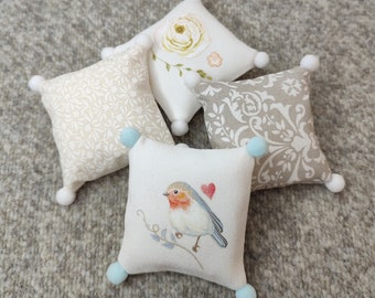 Könnte beinhalten: Vier dekorative Kissen mit verschiedenen Designs. Ein Kissen zeigt ein weißes Blumenmuster, ein anderes ein beigefarbenes Damastmuster, ein drittes ein beige-weißes geometrisches Muster und das vierte einen weißen Hintergrund mit einem blauen Vogel und einem roten Herzen.