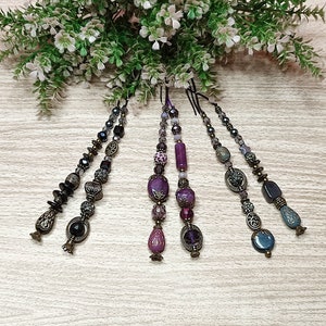 Könnte beinhalten: Fünf Perlen-Schlüsselanhänger mit lila, schwarzen und silbernen Perlen. Die Schlüsselanhänger haben einen Quasten am Ende und hängen an einer grünen, belaubten Pflanze.