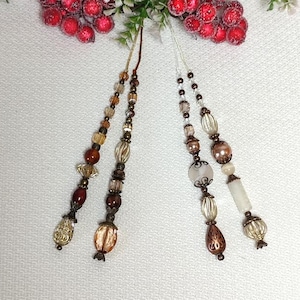 以下が含まれることがあります： 茶色、白、金色のビーズがついた4本のビーズの紐。紐は白い表面にぶら下がっている。