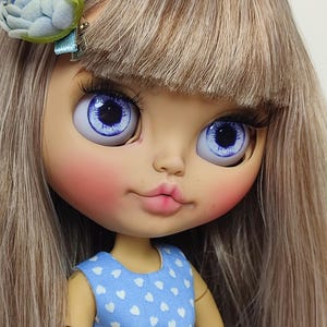 以下が含まれることがあります： 長い明るい茶色の髪と大きな青い目を持つブライス人形。白いハート模様の青いドレスと青い花のヘアアクセサリーを着用しています。人形はバラ色の頬とソフトピンクの唇の色をしています。