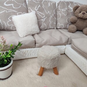K&ouml;nnte beinhalten: Ein wei&szlig;er Hocker aus Kunstfell mit Holzbeinen steht auf einem beigefarbenen Teppich vor einem wei&szlig;en und beigen Ecksofa mit gemusterten Kissen.