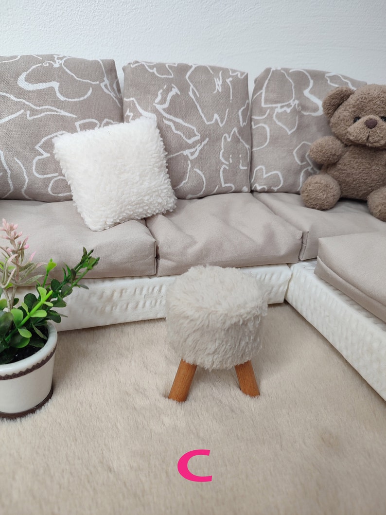 K&ouml;nnte beinhalten: Ein wei&szlig;er Hocker aus Kunstfell mit Holzbeinen steht auf einem beigefarbenen Teppich vor einem wei&szlig;en und beigen Ecksofa mit gemusterten Kissen.