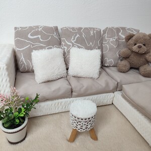 K&ouml;nnte beinhalten: Ein wei&szlig;es, geflochtenes Ecksofa mit beigen Kissen und zwei wei&szlig;en, flauschigen Kissen. Ein kleiner wei&szlig;er Hocker mit einer geh&auml;kelten Oberseite steht vor dem Sofa. Ein brauner Teddyb&auml;r sitzt auf dem Sofa.