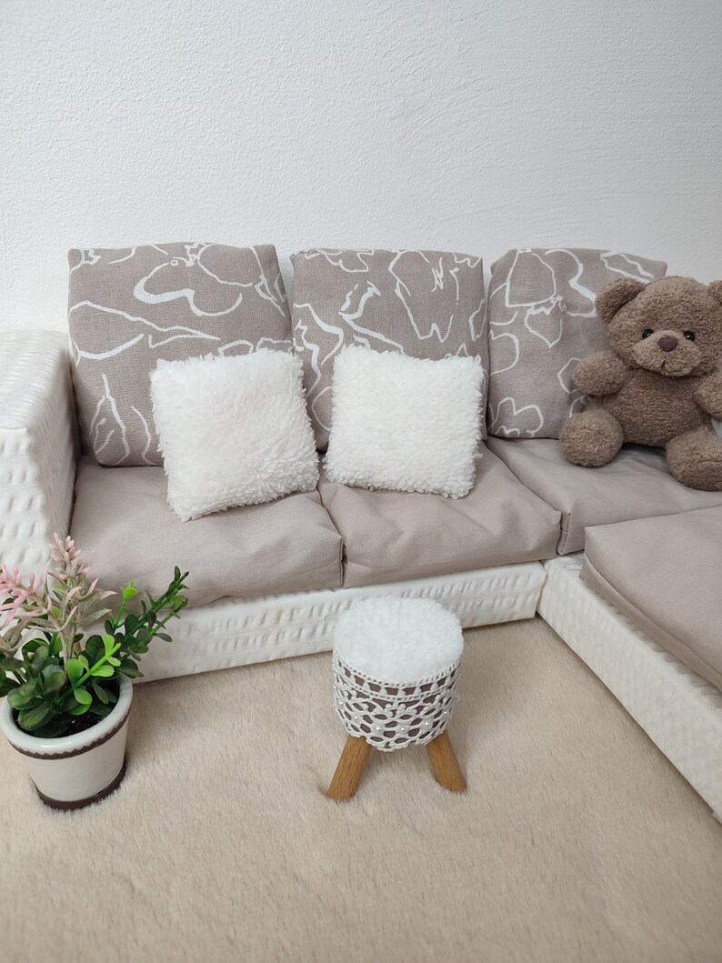 K&ouml;nnte beinhalten: Ein wei&szlig;es, geflochtenes Ecksofa mit beigen Kissen und zwei wei&szlig;en, flauschigen Kissen. Ein kleiner wei&szlig;er Hocker mit einer geh&auml;kelten Oberseite steht vor dem Sofa. Ein brauner Teddyb&auml;r sitzt auf dem Sofa.