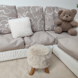 K&ouml;nnte beinhalten: Ein beiges Ecksofa mit wei&szlig;en gemusterten Kissen und einem wei&szlig;en Kissen. Ein brauner Teddyb&auml;r sitzt auf dem Sofa. Ein flauschiger wei&szlig;er Hocker mit Holzbeinen steht auf einem beigefarbenen Teppich vor dem Sofa.