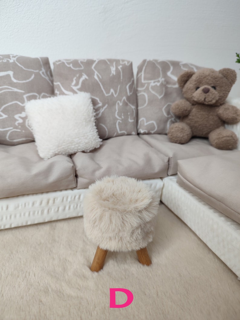 K&ouml;nnte beinhalten: Ein beiges Ecksofa mit wei&szlig;en gemusterten Kissen und einem wei&szlig;en Kissen. Ein brauner Teddyb&auml;r sitzt auf dem Sofa. Ein flauschiger wei&szlig;er Hocker mit Holzbeinen steht auf einem beigefarbenen Teppich vor dem Sofa.