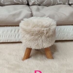 K&ouml;nnte beinhalten: Ein kleiner, flauschiger Hocker in Cremefarbe mit Holzbeinen. Der Hocker steht auf einem cremefarbenen Teppich. Der Buchstabe "D" ist in Pink auf den Teppich gedruckt.