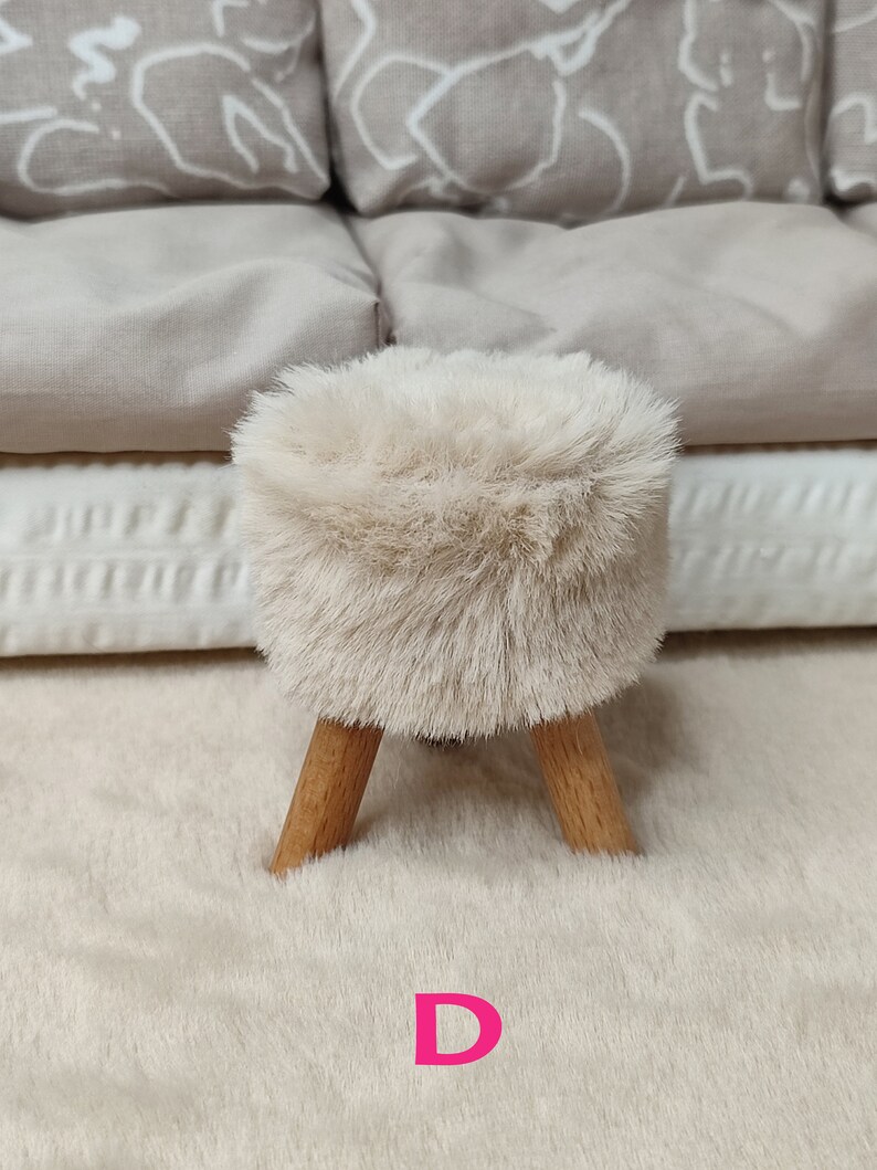 K&ouml;nnte beinhalten: Ein kleiner, flauschiger Hocker in Cremefarbe mit Holzbeinen. Der Hocker steht auf einem cremefarbenen Teppich. Der Buchstabe "D" ist in Pink auf den Teppich gedruckt.