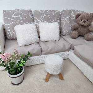 K&ouml;nnte beinhalten: Ein wei&szlig;es Sofa mit drei grauen gemusterten Kissen und zwei wei&szlig;en flauschigen Kissen. Ein Teddyb&auml;r sitzt auf dem Sofa. Ein wei&szlig;er flauschiger Hocker mit Holzbeinen steht auf einem wei&szlig;en flauschigen Teppich. Eine Topfpflanze mit rosa Bl&uuml;ten steht auf dem Teppich. Der Buchstabe "A" ist in rosa auf dem Teppich geschrieben.
