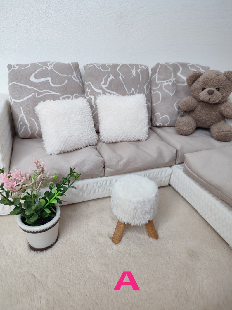 K&ouml;nnte beinhalten: Ein wei&szlig;es Sofa mit drei grauen gemusterten Kissen und zwei wei&szlig;en flauschigen Kissen. Ein Teddyb&auml;r sitzt auf dem Sofa. Ein wei&szlig;er flauschiger Hocker mit Holzbeinen steht auf einem wei&szlig;en flauschigen Teppich. Eine Topfpflanze mit rosa Bl&uuml;ten steht auf dem Teppich. Der Buchstabe "A" ist in rosa auf dem Teppich geschrieben.